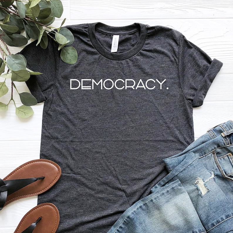 Puede incluir: Una camiseta gris oscuro con la palabra "DEMOCRACY." impresa en blanco en la parte delantera.