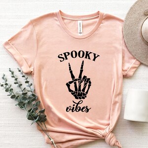 May include: A light pink t-shirt with the words "SPOOKY vibes" printed in black. The design features a black skeleton hand making a peace sign. The shirt is folded and has a rolled sleeve.