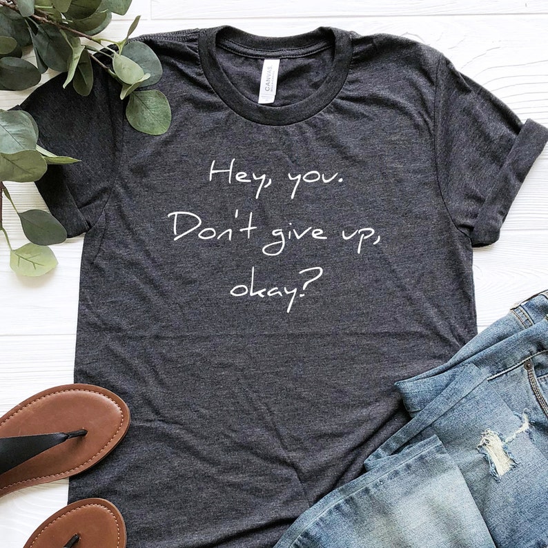 May include: A dark heather grey t-shirt with the text "Hey you. Don't give up, okay?" written in white.