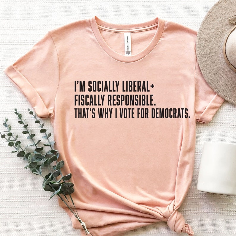 May include: A peach-colored t-shirt with the text "I'M SOCIALLY LIBERAL+ FISCALLY RESPONSIBLE. THAT'S WHY I VOTE FOR DEMOCRATS."
