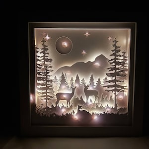 May include: A white wooden shadow box with a cut-out design of a forest scene with two deer, a mountain, trees, and a moon. The shadow box is illuminated with white LED lights.