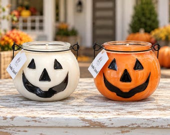 May include: Two glass jar candles shaped like jack-o'-lanterns, one white and one orange, each featuring a black painted face, a black wire handle, and a white rectangular tag labeled Pumpkin Caramel hanging from the side.