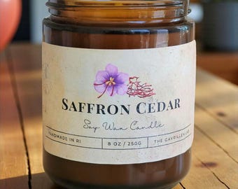 May include: A brown glass jar candle with a label that reads "Saffron Cedar Soy Wax Candle." The label features a purple flower illustration and the text "Handmade in RI." The candle is 8 oz / 250g.