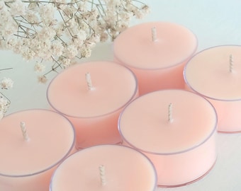 Może przedstawiać: Sześć okrągłych, bladoróżowych świec typu tealight z białymi knotami ułożonych w grupie. Świece są w przezroczystych plastikowych kubeczkach. Białe kwiaty w tle.