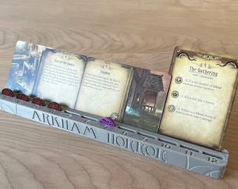 May include: A gray 3D-printed plastic card holder for Arkham Horror featuring slots for game cards and numbered spaces from 1 to 12. The holder contains several game cards, including The Gathering, and small red and purple circular tokens.