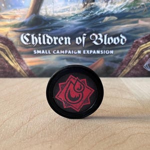 Puede incluir: Una ficha de juego circular negra con un icono rojo en forma de estrella y un diseño de gota en el centro. La ficha está colocada frente a una caja de juego etiquetada como Children of Blood Small Campaign Expansion con una ilustración.