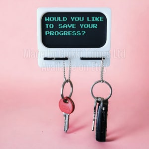 Peut inclure: Porte-clés blanc en forme d'écran d'ordinateur rétro avec le texte "WOULD YOU LIKE TO SAVE YOUR PROGRESS?" et les mots "Made by Chikki Things Ltd Available on Etsy". Deux porte-clés sont suspendus au support, l'un avec une clé rouge et l'autre avec un jeu de clés.