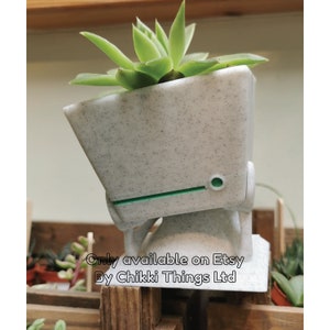 May include: A white and grey speckled planter shaped like a robot head with a green stripe and a small green circle on the side. The planter is holding a succulent plant. The text "Only available on Etsy By Chikki Things Ltd" is visible below the planter.
