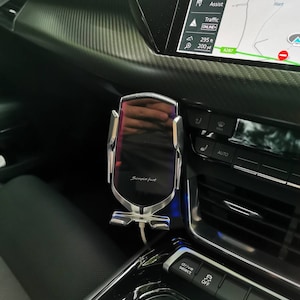 Puede incluir: Un soporte para teléfono de coche plateado y negro con el texto "Simple fast" está montado en el salpicadero de un coche. Un cable de carga blanco está conectado al soporte. La pantalla de infoentretenimiento del coche muestra un mapa e información de navegación.