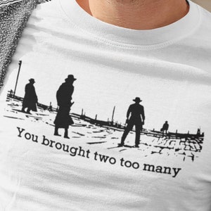 May include: White t-shirt with a black silhouette of three figures walking away from the viewer. The text "You brought two too many" is printed below the figures.
