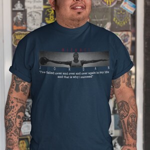 Op de afbeelding: Een marineblauw t-shirt met een zwart-wit afbeelding van Michael Jordan die basketbal speelt. De tekst "MICHAEL JORDAN" is in rood boven de afbeelding gedrukt. De quote "Ik ben keer op keer gefaald in mijn leven en dat is waarom ik succesvol ben" is in wit onder de afbeelding gedrukt.