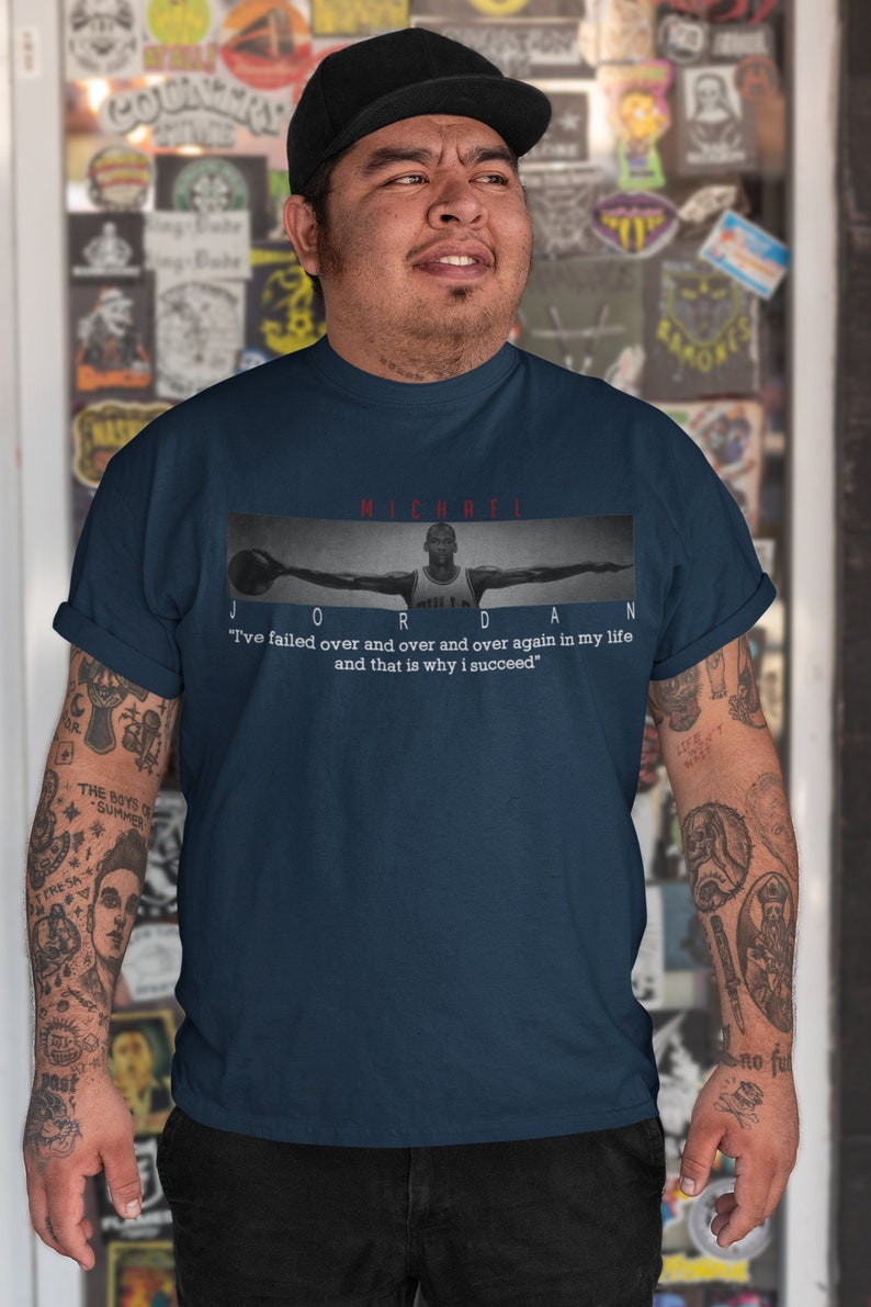 Op de afbeelding: Een marineblauw t-shirt met een zwart-wit afbeelding van Michael Jordan die basketbal speelt. De tekst "MICHAEL JORDAN" is in rood boven de afbeelding gedrukt. De quote "Ik ben keer op keer gefaald in mijn leven en dat is waarom ik succesvol ben" is in wit onder de afbeelding gedrukt.