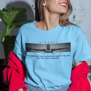 Op de afbeelding: Lichtblauw T-shirt met een zwart-witte afbeelding van Michael Jordan met de tekst "MICHAEL JORDAN" en de quote "Ik ben keer op keer gefaald en dat is waarom ik succesvol ben."