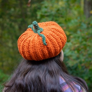 May include: A crocheted pumpkin hat in orange with a green stem. The hat is worn on a person's head with long brown hair.