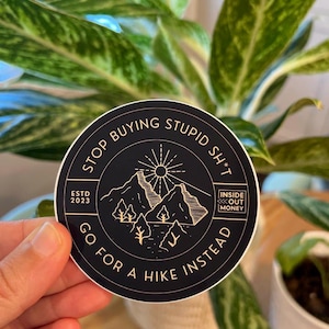 May include: A black and gold round sticker with the text "STOP BUYING STUPID SH*T" and "GO FOR A HIKE INSTEAD". The sticker also features a mountain scene with a sun and the text "INSIDE OUT MONEY" and "ESTD 2023".