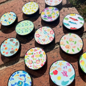 May include: A collection of 12 colourful phone grips with various summer-themed designs. The designs include lemons, pineapples, watermelon, peaches, seashells, and more. The phone grips are all round and have a black base.