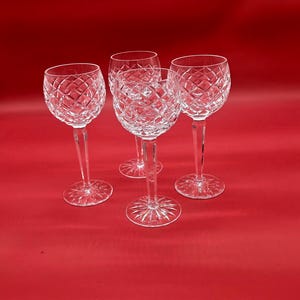 May include: Four clear crystal wine glasses with diamond-cut patterns. The glasses have long stems and round bases, set against a solid red background. The wine glasses are designed for serving wine.