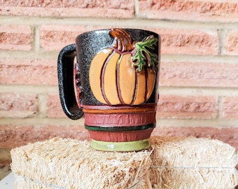 May include: A ceramic mug with a black textured finish featuring a raised, hand-painted orange pumpkin with a green leaf. The base of the mug has horizontal bands in dark red, green, and light green, with a sturdy black handle on the side.