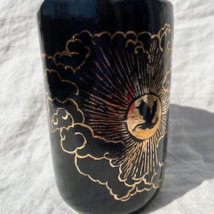 May include: Black ceramic tumbler with a gold sun and cloud design. The sun is centered with radiating lines, and the clouds are outlined in gold. The tumbler has a glossy finish and a slightly tapered shape.
