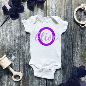 Könnte beinhalten: Weißer Baby-Strampler mit einem lila Kreis und dem Namen "Olive" in lila Schrift.