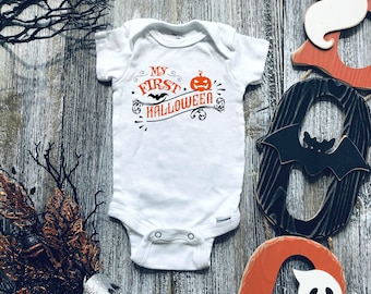 Puede incluir: Un body blanco para bebé con el texto "My First Halloween" en negro con un gráfico de calabaza naranja. El body está sobre un fondo de madera con decoraciones de Halloween.