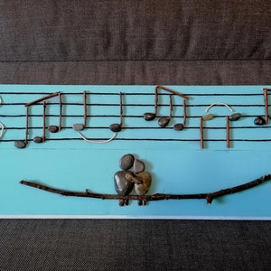 Può includere: Un pannello di legno blu con un pentagramma dipinto su di esso. Il pentagramma è decorato con piccole pietre nere e ramoscelli per rappresentare le note musicali. Un ramo di ramoscelli con due piccoli uccelli di pietra si trova sotto il pentagramma.
