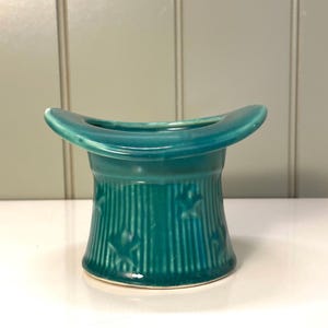 May include: A teal ceramic top hat-shaped container. The container has vertical lines and star-shaped cutouts. The hat has a wide brim and a cylindrical body. The container is sitting on a white surface.