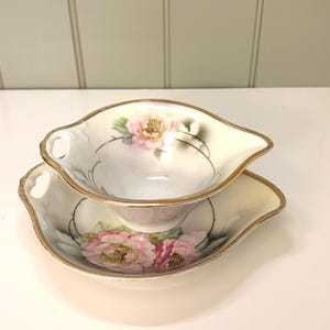 May include: A cream-colored, vintage gravy boat and underplate set. The set features a floral design with pink and yellow flowers and gold trim. The gravy boat has a handle on each side.
