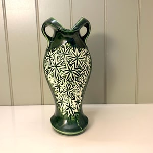 May include: A dark green ceramic vase with a flared base and two handles. The vase features a white starburst pattern over the dark green glaze. The vase is a decorative item.