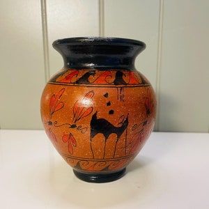 May include: A small, brown ceramic vase with a black rim and a black and red painted design. The design features a stylized animal and geometric patterns.