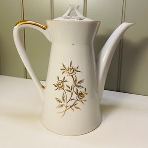 May include: White ceramic coffee pot with a gold trim and floral design. The pot has a lid with a gold handle.