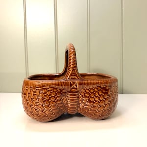 May include: A brown ceramic basket-shaped container with a handle. The basket has a woven texture and two compartments. The handle is arched and also has a woven texture. The container is sitting on a white surface.
