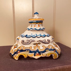 May include: A white ceramic inkwell with a blue and yellow floral design. The inkwell has a domed lid and a scalloped base.