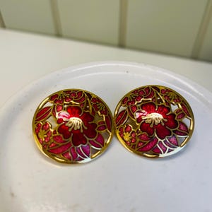 May include: A pair of gold-toned, round earrings with a floral design. The earrings feature a red and pink flower with gold accents. The floral design is set against a white background. The earrings are on a white surface.