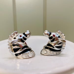May include: A pair of silver tone zebra head earrings with black and white stripes and red rhinestone eyes. The earrings are adorned with clear rhinestones.