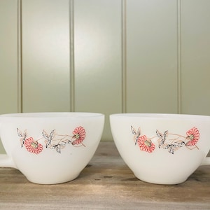 May include: Two white ceramic teacups with a floral design. The cups have a red and black floral pattern with a vine design. The cups are on a wooden surface.