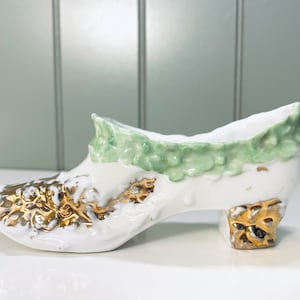 May include: A white porcelain shoe with gold and green accents. The shoe has a green floral design around the top and gold floral designs on the sides and heel.
