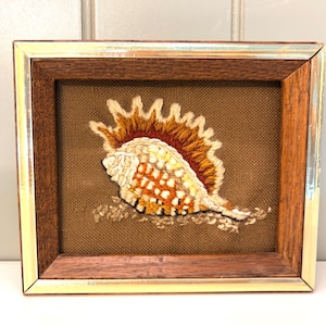 May include: A framed embroidery piece featuring a detailed seashell design in shades of orange, brown, and cream. The shell is stitched onto a textured brown fabric background and enclosed within a wooden frame with a metallic gold-toned border.