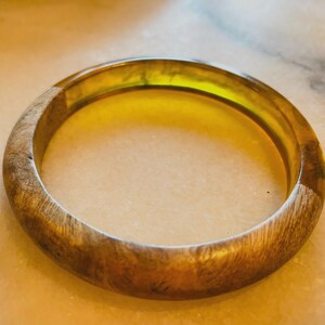 May include: A circular bangle bracelet featuring a combination of natural brown wood grain and a translucent, bright yellow resin section. The smooth, polished surface displays a distinct contrast between the organic wood texture and the clear resin.