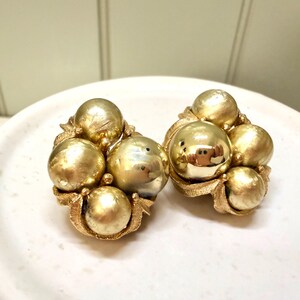 May include: A pair of vintage gold-tone clip-on earrings. Each earring features a cluster of six textured, spherical orbs. The orbs are arranged within a decorative, textured gold-tone frame. The earrings are displayed on a white, round surface.