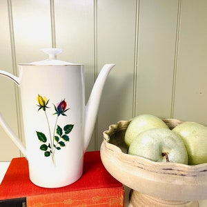 May include: White ceramic coffee pot with a gold trim and a floral design featuring yellow, red, and green roses. The pot is sitting on a red book.