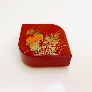 May include: A red lacquered box with a curved edge. The top is decorated with gold and orange floral designs. The box has a glossy finish and is set against a white background.