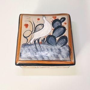 May include: A square ceramic box with a hand-painted design of a white bird with black accents standing in front of a cactus. The box has a brown border and a white background.