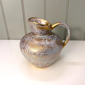 May include: A decorative ceramic pitcher with a gold and white textured finish. The pitcher has a rounded body, a handle, and a spout. The gold accents the rim and handle, adding a touch of elegance to the piece. The pitcher is approximately 8 inches tall.
