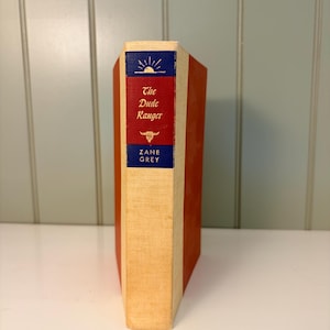 May include: A hardcover book titled "The Dude Ranger" by Zane Grey. The book has a red spine and a cream-colored front and back cover. The book's spine features a blue band with a sunburst design and the author's name.
