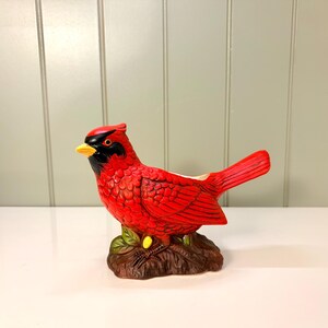 May include: A ceramic cardinal bird figurine featuring vibrant red feathers, a black mask around the eyes, and a yellow beak. The bird is perched on a textured brown branch base with green leaves, and the top of the bird has an open cavity.