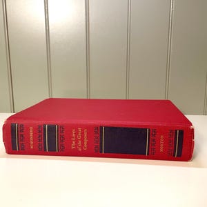 May include: A red hardcover book titled "The Lives of the Great Composers" by  the author,  "M.D. Calvocoressi" with the publisher "W.W. Norton & Company, Inc."