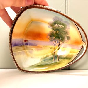 May include: A hand-painted, vintage porcelain dish with a landscape scene. The dish features a sunset with trees, a small house, and a brown rim. The dish is held by a hand.