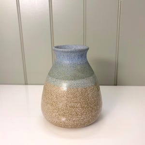 May include: A handcrafted ceramic vase with a speckled, textured surface. The vase features a color gradient, transitioning from a light blue rim to a green midsection and a tan base. The vase has a rounded body and a narrow neck.