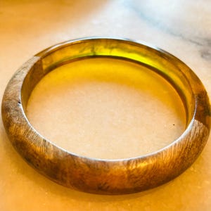 May include: A round bangle bracelet with a wood-grain texture and a translucent amber-colored center. The bracelet has a smooth, polished finish, and the wood grain is visible on the outer edges. The overall design is simple and elegant.